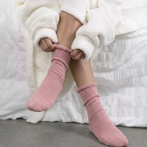 Cotton sleep socks in dusty pink ribbed knit, featuring a rolled cuff. Worn on feet while seated on a cozy bed.