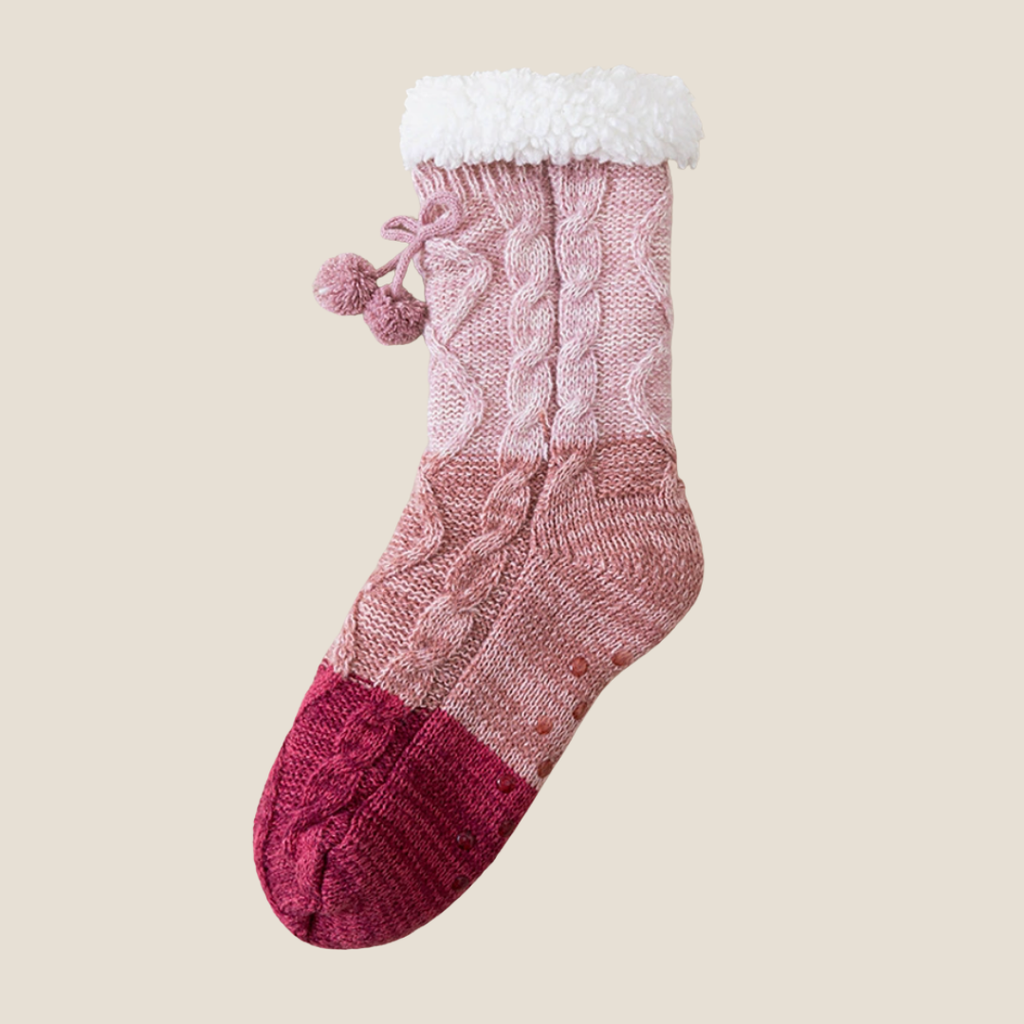 Crochet slipper socks in pink, featuring pom-pom accents and a knit cuff. Shown as a single flat-lay on a neutral backdrop.