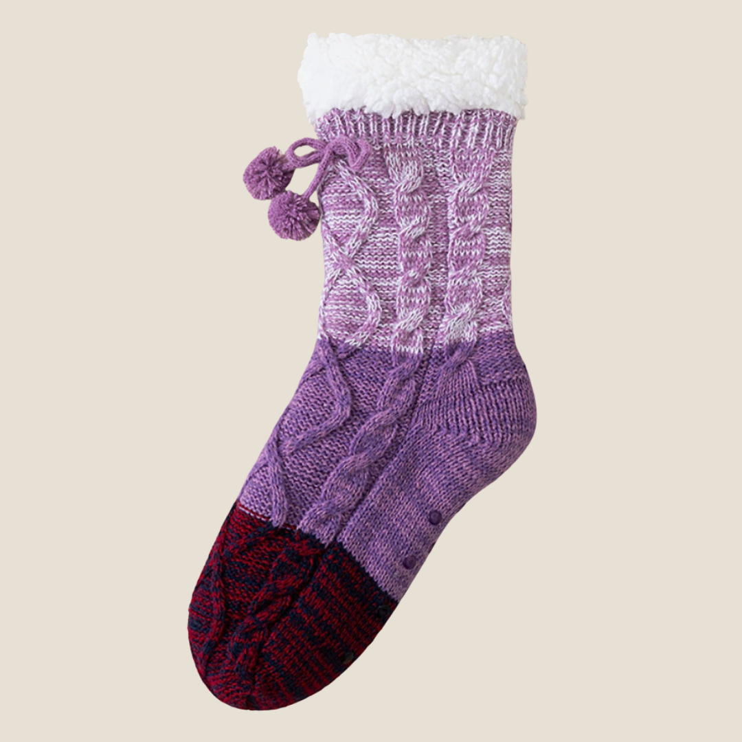 Crochet slipper socks in purple colorblock knit, featuring pom-pom ties and a plush cuff. Displayed flat on a light surface.