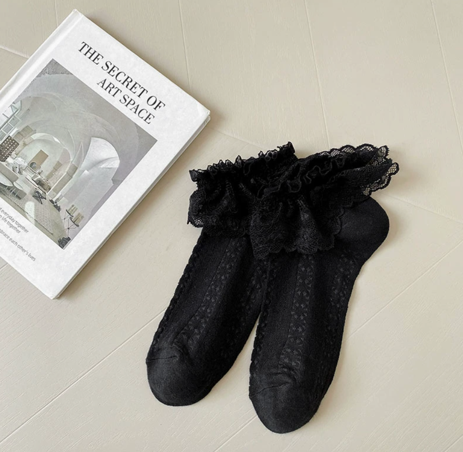 Lace ankle socks in black, featuring a frilled lace trim. Shown as a flat-lay pair beside a book.