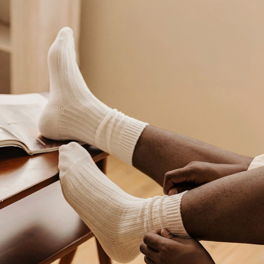 Men's sleep socks in cream ribbed cotton, featuring a soft relaxed cuff. Worn on feet in a warm indoor setting.