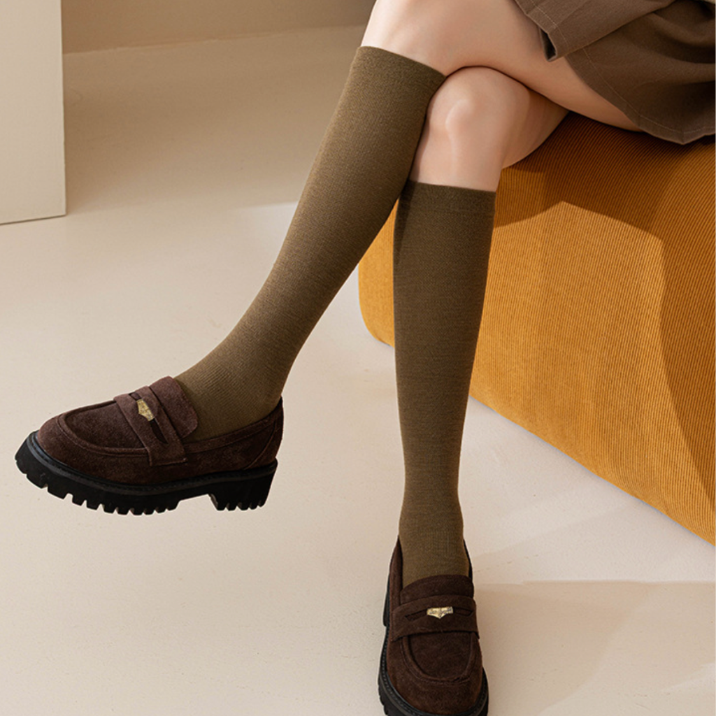 Merino wool knee socks in olive green ribbed knit, featuring a soft cuffed top. Worn on legs with brown loafers in a cozy seated look.