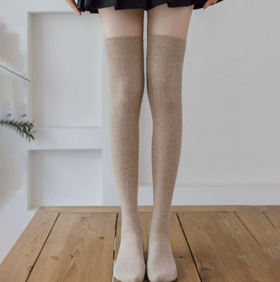 Over knee socks in cream ribbed knit, featuring a thigh-high length. Worn standing on a wooden floor.