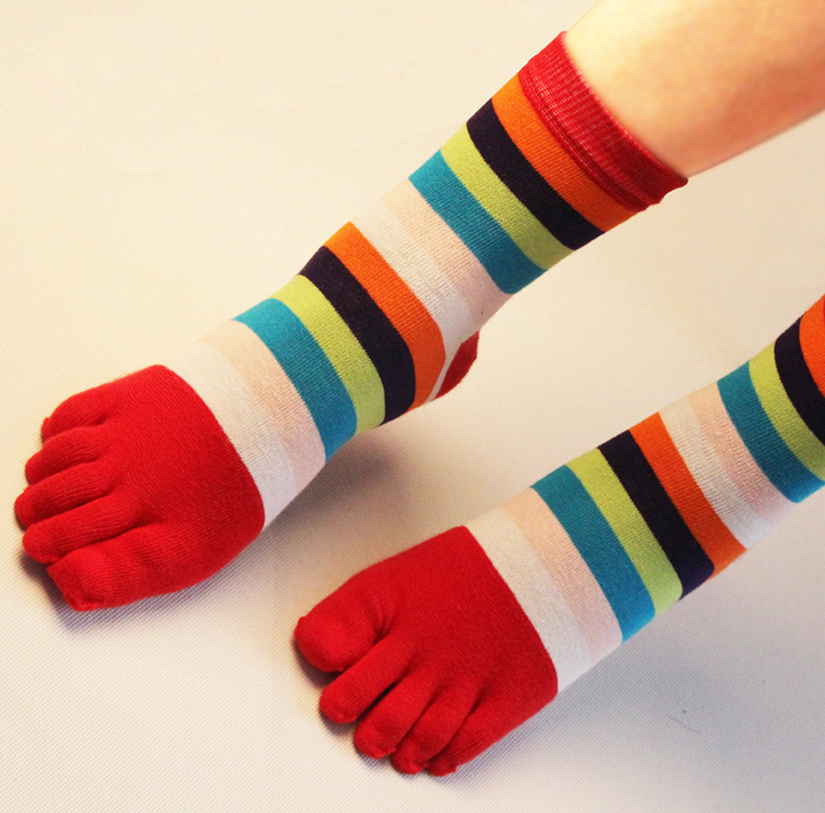 Rainbow toe socks in multicolor stripes with red toes, featuring individual toe pockets. Worn on crossed legs.