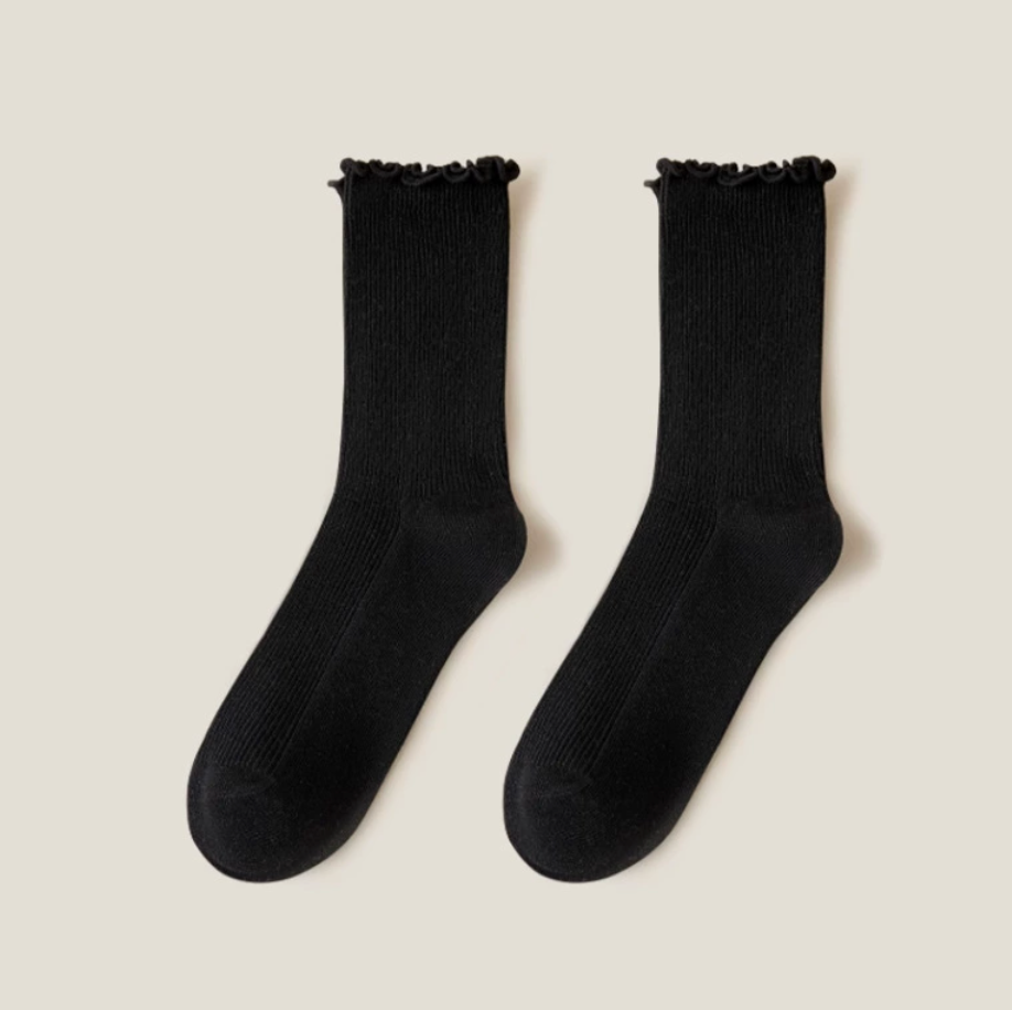 Ruffle crew socks in black ribbed knit, featuring a frilled ruffle cuff. Shown as a flat-lay pair on a light surface.