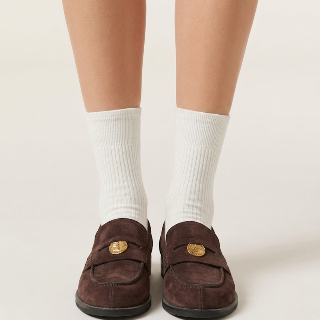 Socks for loafers women in white ribbed cotton, worn with brown penny loafers. Front view of both feet on a plain backdrop.