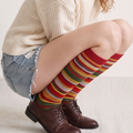 Striped knee high socks in multicolor rainbow stripes, featuring a retro banded knit. Worn seated with denim shorts and brown boots.