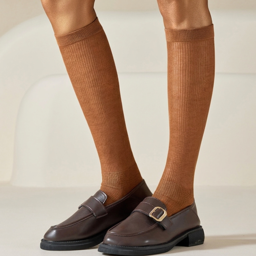 Thin socks for loafers in caramel brown ribbed knit, styled on the legs with brown buckle loafers, side view.