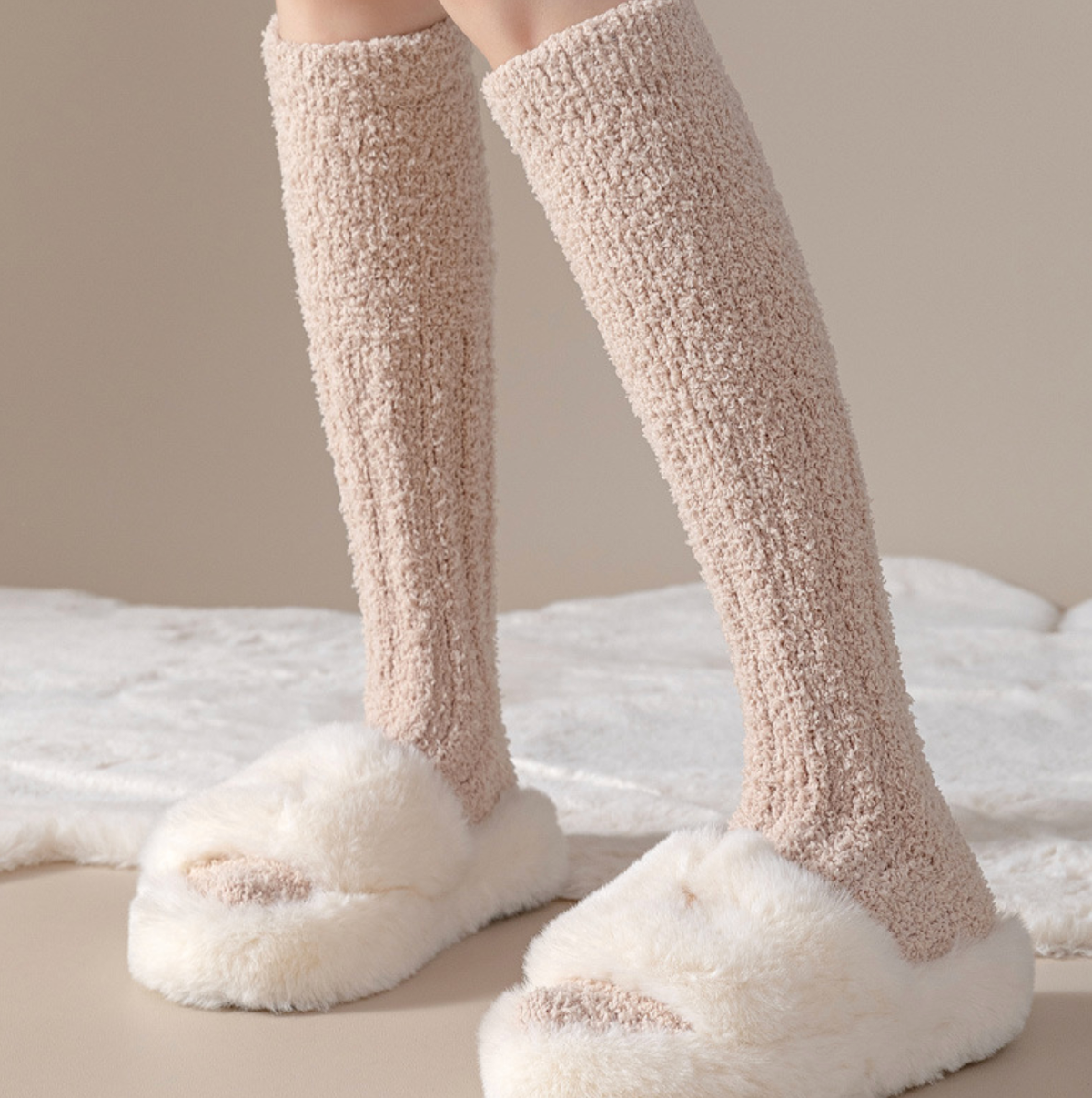 Warm knee high socks in cream fuzzy plush, featuring a fluffy cozy knit. Worn with fluffy slippers in a warm setting.