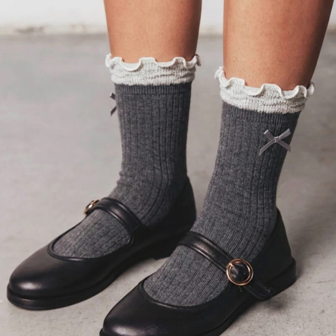 Women's cute socks in gray ribbed knit with a white lace ruffle and bow, worn with black Mary Jane shoes, front view.