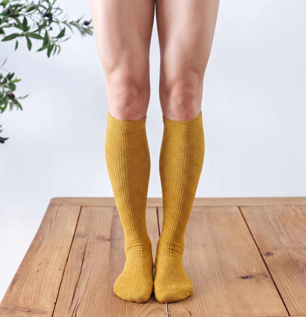 Women's knee socks in mustard yellow ribbed knit, featuring a knee-high leg. Worn standing on a wooden floor.