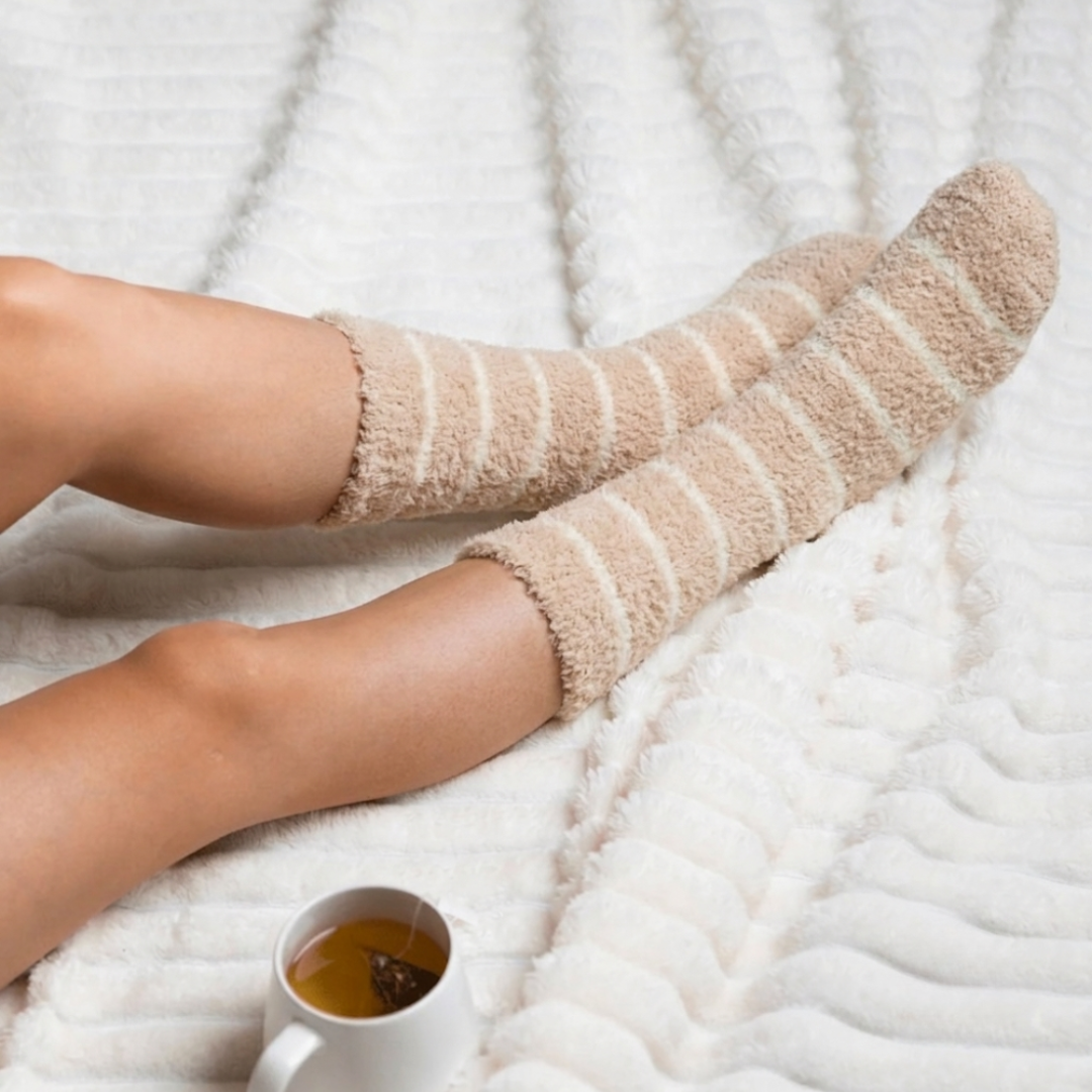 Women's sleep socks in oatmeal and cream plush, featuring fuzzy striped bands. Worn on legs on a knit blanket beside a warm drink.